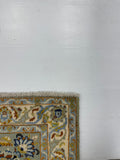 A close-up of the corner of a patterned area rug with fringe, shown on a plain white floor. The rug features a cream base with blue, gold, and brown floral motifs.