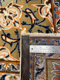 Close-up of the corner of a patterned area rug with a metal ruler and leather sample strip placed to show dimensions and texture. The rug has an ornate floral and scroll motif in beige, blue, orange, brown, and cream.