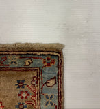 A close-up corner view of a patterned area rug with a beige field, blue border, and red accents, shown against a white textured wall.