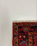 A close-up corner of a patterned rug on a white textured surface, showing red, black, blue, and beige woven details with cream fringe.