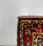 A close-up corner of a patterned area rug with cream, red, navy, and orange floral motifs, shown against a plain white wall.