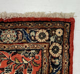 A close-up of a patterned area rug corner with a red border and cream fringe, laid against a large white board on a gray carpeted floor.