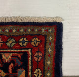 A close-up corner of a patterned area rug with cream fringe, showing a red field, dark navy border, and floral motifs in beige, green, and blue, photographed against a plain white surface.