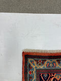 A close-up corner view of a patterned area rug laid on a white surface, showing the fringe edge, rust-red border, and blue, red, beige, and navy woven design.