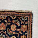 A close-up corner of a patterned rug with dark navy, rust orange, beige, and black floral motifs, shown on a white board over a gray carpet.