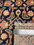 Close-up of a patterned rug with a stainless steel ruler placed along the edge to show scale. The rug has a dark navy blue base with intricate floral and paisley motifs in orange, beige, and muted green, and a fringed edge is visible.