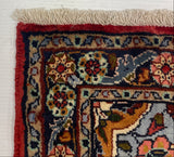 A close-up corner of a patterned area rug laid on a white surface, showing cream fringe, a red border, and a multicolored floral and geometric field in navy, red, beige, and orange tones.