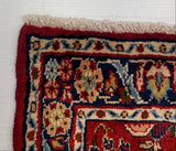 A close-up corner view of a patterned area rug laid on a white surface, with a gray carpet visible at the top. The rug has a red border, cream fringe, and a multicolor floral and geometric pattern in blue, beige, and red.