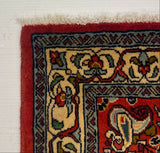 A close-up of a patterned rug corner showing a red border, cream and red central field, and dark blue geometric accents, photographed against a white board with carpet visible below.