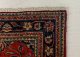A close-up of a patterned area rug corner placed on a white wooden board, with gray carpet visible around the edges. The rug has a red, navy, and beige floral motif.