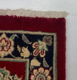 A close-up corner of a patterned area rug with a deep red field, navy border, and cream floral motifs, photographed against a plain white backdrop.