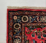 A close-up corner of a patterned area rug with a red field and ornate dark blue, beige, and green floral border, photographed against a white floor with a gray carpeted surface above.