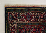 A close-up of a patterned area rug corner with cream fringe, laid against a large white board on a gray carpeted floor.