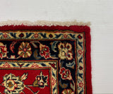 A close-up corner of a patterned area rug with a red field and dark navy border, laid against a large white board on a gray carpeted floor.