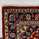 A corner view of a traditional patterned rug laid on a white board over a gray carpet. The rug has a deep red border, navy blue field, and cream floral motifs with small red, green, and gold accents.