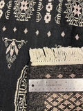 Close-up of a woven rug with cream fringe, a dark charcoal/black base, and light pink and cream geometric patterns, shown with a metal ruler for scale.
