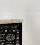 A close-up of a black woven rug with cream geometric patterns and fringe, laid on a white surface against a gray textured background.