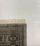 A close-up corner view of a patterned area rug with cream fringe, laid on a large white board over a gray carpeted floor.