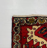 A close-up corner view of a patterned area rug laid on a white board, with carpet visible above the board. The rug has a red field, cream central motif, and dark navy floral border.