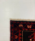 A close-up of a patterned area rug corner placed on a white board over a gray carpet. The rug has a dark navy base with red, orange, and cream floral motifs and a cream fringe.