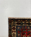 A close-up of a patterned area rug corner with a dark navy blue field, red and green motifs, and a beige border, laid on top of a large white board over a gray carpet.