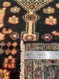 Close-up of a patterned area rug with a dark navy or black base, cream, orange, and green geometric motifs, a visible fringe edge, and a stainless steel ruler placed along the side for scale.