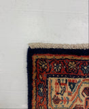 A close-up corner view of a patterned area rug laid on a white surface. The rug has a dark navy blue border, a red and beige central field, and cream fringe along the top edge.