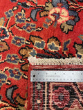 A close-up of a patterned area rug with a red field, cream fringe, and a stainless steel ruler placed vertically to show scale. The rug features blue, beige, and black floral motifs.