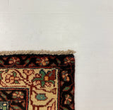 A corner view of a patterned area rug with a dark red border and beige field, partially shown on top of a large white board against a gray carpeted floor.