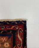 A close-up corner view of a patterned area rug with a cream frise, dark navy border, and red field, photographed on a white board over a gray carpeted floor.