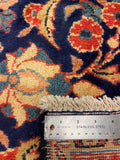 Close-up of a patterned area rug with cream, orange, blue, and navy floral motifs, showing the fringed edge and a stainless steel ruler for scale.