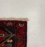A close-up corner of a patterned rug with red, black, beige, and blue motifs, placed on a large white board over a gray floor.