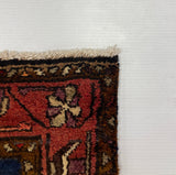 A close-up corner of a patterned area rug with red, brown, blue, and orange motifs, placed against a large white board on a gray carpeted floor.