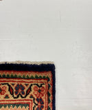 A close-up of the corner of a patterned area rug on a white board, with gray carpet visible around it. The rug has a dark navy border and a cream, red, and blue floral pattern.