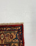 A corner view of a traditional patterned rug with red, beige, and blue details, laid on top of a large white board against a gray carpeted floor.