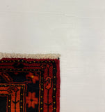 A close-up of a patterned rug corner showing a dark navy and red border with cream fringe, placed against a large white board on a gray carpeted floor.