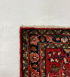 A red patterned area rug with dark borders and a frayed edge, partially visible on a gray carpet next to a large white board.