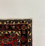 A folded area rug with a traditional pattern in red, navy, and beige, partially visible on a gray carpet next to a white board.