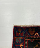 A close-up of a patterned area rug corner placed on a large white board, photographed on a gray carpeted floor. The rug has a dark navy base with red, blue, beige, and orange geometric motifs.