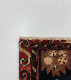 A folded area rug with a dark navy blue field, red border, and beige floral motifs, partially covered by a large white cardboard sheet on a gray carpeted floor.