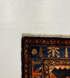 A close-up of a patterned rug corner placed on a white board over a gray carpeted floor. The rug shows a dark navy and black border with red, orange, and beige patterned sections.