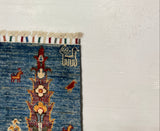 A close-up of a blue patterned rug corner with multicolor floral motifs and fringe, laid over a large white board on a gray carpeted floor.