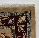 Close-up of a patterned area rug corner showing a beige border, dark red field, and cream fringe, laid against a white backdrop.