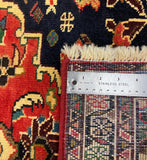 Close-up of a patterned rug corner with a stainless steel ruler placed along the edge to show the pile height and weave. The rug has a dark navy field with cream, red, orange, and beige floral motifs and a multicolor geometric border.