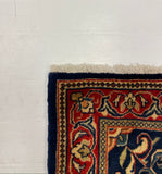 A close-up corner of a patterned area rug with cream fringe, featuring a navy blue border and red, beige, and gold floral motifs, shown on a white floor against a gray carpet.