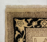 Close-up corner of a woven rug showing a beige and black floral pattern with a fringed edge, photographed against a white wall.