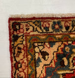 A corner view of a traditional patterned area rug with red, beige, and blue motifs, laid on top of a large white board against a gray carpeted floor.
