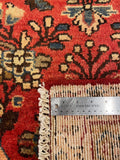Close-up of a patterned area rug corner with a metal ruler placed along the edge to show the pile height and weave detail. The rug features a red field with cream, beige, and dark brown floral motifs.