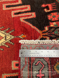 Close-up of a patterned rug with a metal ruler placed vertically to show the weave and scale. The rug features a red field with cream, black, and gray geometric and floral motifs.