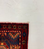 A close-up of a patterned area rug corner placed on a white board over a gray carpeted floor. The rug shows a deep red border and a blue and red central field with geometric and floral motifs.